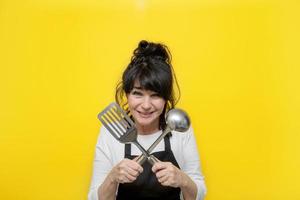 laughing senior woman in black apron on yellow background holding crossed scoop and shovel, seniors activity concept, tasty food and cooking photo