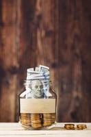 A glass jar with coins and a hundred-dollar bill, a sticker with free space for text, on a wooden table. photo