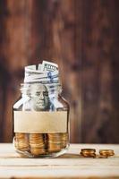 Glass jar with coins and dollars, with a sticker with free space for text, on a wooden background. photo