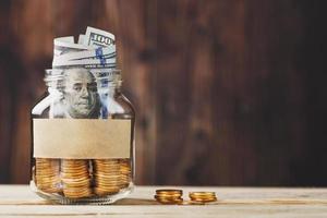 Glass jar with coins and dollars, with a sticker with free space for text, on a wooden background. photo