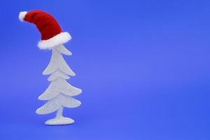 White shiny Christmas tree with Santa Claus hat on its head on blue background. New Year. Copy space photo