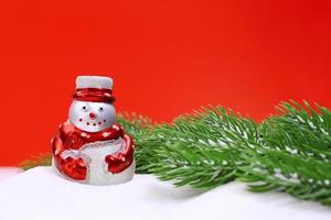 Snowman in the snow and green spruce branches on red background. Christmas, New Year. Copy space photo