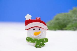 Snowman with a red cap and a green scarf on a snowy background and green trees. Christmas, New Year. Copy space photo