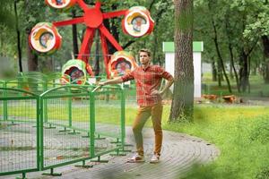 a young man in a plaid shirt is standing in a park with a phone in his hands, behind him is an attraction photo