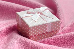 A small gift box in pink with a small bow lies on a blanket of soft and furry light pink fleece fabric with a lot of relief folds. Packing for a gift to your lovely girlfriend photo