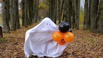 A child in sheets with cutout for eyes like a ghost costume dancing in an autumn forest scares and terrifies. A kind little funny ghost. Halloween Party, slow motion 4k video