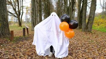 A child in sheets with cutout for eyes like a ghost costume dancing in an autumn forest scares and terrifies. A kind little funny ghost. Halloween Party, slow motion 4k video