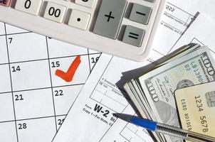 declaración de impuestos y salarios w-2 en blanco con tarjeta de crédito en billetes de dólar, calculadora y bolígrafo en la página del calendario con el 15 de abril marcado foto