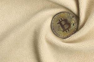 The golden bitcoin lies on a blanket made of soft and fluffy light orange fleece fabric with a large number of relief folds. The shape of the folds resembles a fan from a video card cooler photo