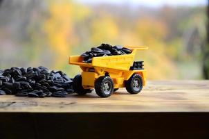 A small yellow toy truck is loaded with sunflower seeds next to a small pile of sunflower seeds. A car on a wooden surface against a background of autumn forest. Transportation of sunflower seeds photo