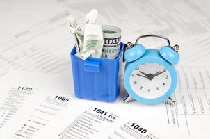 Many american tax blank forms with blue alarm clock and crumpled hundred dollar bill in trash bin photo