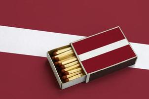 Latvia flag  is shown in an open matchbox, which is filled with matches and lies on a large flag photo