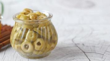 Small glass jar full of sliced green olives with cup of oil in background video