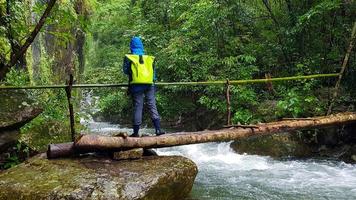 Backpacker or traveler wear blue raincoat with green backpack, stand and cross on wooden bridge with waterfall and tree background in tropical deep jungle or forest at Chiang Mai, Thailand. Adventure photo