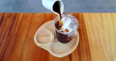 Pouring black or dark coffee in plastic glass of ice with fresh coconut on brown wooden table at cafe. Cold drinking and refreshment. Iced coconut coffee menu. photo