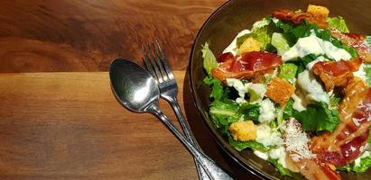 Fresh Caesar salad with vegetable, fried bacon, crispy bread, spoon and fork on brown wooden background or table with copy space. Healthy food topping with cream sauce. Good lifestyle eating concept. photo