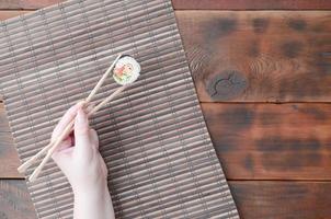 A hand with chopsticks holds a sushi roll on a bamboo straw serwing mat background. Traditional Asian food photo