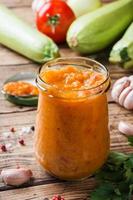 Homemade caviar from zucchini tomatoes and onions in a glass jar on a wooden background. Homemade production canning, canned stewed vegetable. photo