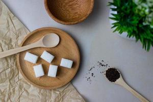 Sugar cubes in a wooden plate and spoon with tea feaves on the kitchen table with utensils and green plant. Top view photo