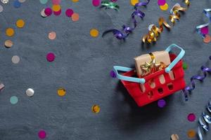 Shopping basket with gift box and festive tinsel flat lay on dark background. Top view photo