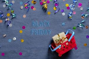 Black Friday tetx with shopping basket and festive tinsel flat lay on dark background. Top view photo