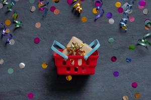 Shopping basket with gift box and festive tinsel flat lay on dark background. Top view photo