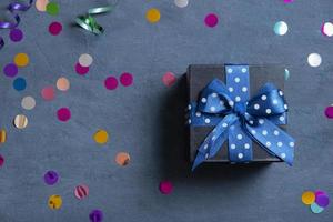 Black gift box with bow and holiday tinsel flat lay on dark cement background. Top view photo