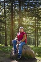 A young man in a sleeveless red jacket is sitting on a huge stone in a pine forest in spring. The magical light from the sun's rays falls behind the boy. photo