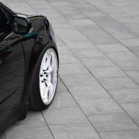 Diagonal view of a black glossy car with white wheels, which stands on a square of gray tiles photo