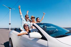 Nothing but friends and road ahead. Group of young happy people enjoying road trip in their white convertible and raising their arms up photo