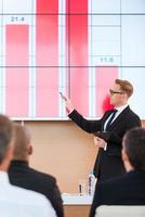 Presentation in conference hall. Confident young man in formalwear pointing projection screen with graph on it while making presentation in conference hall with people on the foreground photo