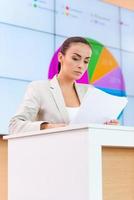 Confident public speaker. Confident young woman in formalwear standing at the tribune and looking at paper while making a presentation with projection screen in the background photo