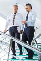 Discussing business contract. Two confident businessmen discussing something and looking at paper while moving down by staircase photo