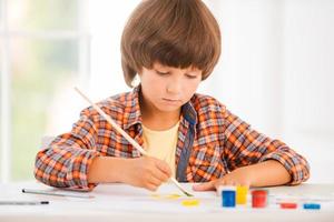 pequeño artista niño concentrado relajándose mientras pinta con acuarelas sentado en la mesa foto