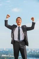 I did it Happy young man in formalwear keeping arms raised and expressing positivity while standing outdoors with cityscape in the background photo