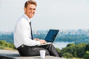 tener libertad para trabajar en todas partes. joven confiado con camisa y corbata trabajando en una laptop y sonriendo mientras se sienta al aire libre con el paisaje urbano en el fondo foto