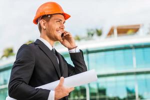 Confident architect. Handsome young man in hardhat holding blueprint and talking on the mobile phone while standing outdoors and against building structure