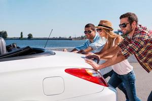 Never desponding. Three young people pushing their broken convertible and smiling photo