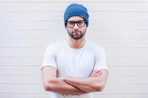 joven y creativo. apuesto joven con anteojos manteniendo los brazos cruzados y mirando a la cámara mientras está de pie contra la pared de ladrillo foto