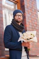 The freshest roses for her. Handsome young man in smart casual wear holding bouquet of flowers and smiling while standing at the street photo