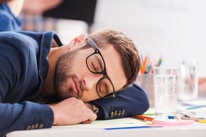 durmiendo en el trabajo. joven hombre de negocios cansado durmiendo sentado en su lugar de trabajo e inclinando la cabeza en el escritorio foto