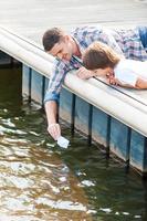 Having fun together. Cheerful father and son playing with paper boat while lying down on the quayside together photo