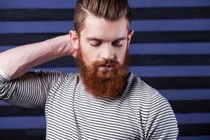 Casual and cool. Side view of handsome young bearded man holding hand on head while standing against stripped background photo