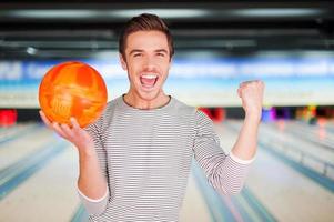 el campeón de bolos. joven alegre sosteniendo una bola de boliche y manteniendo los brazos levantados mientras se enfrenta a las boleras foto