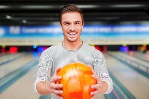 confiado y creativo. joven alegre estirando una bola de bolos y sonriendo mientras está de pie contra las boleras foto