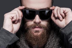 Always in style. Portrait of handsome young bearded man adjusting his sunglasses and looking at camera while standing against grey background photo