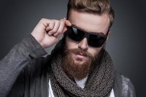 Confident in his perfect style. Portrait of handsome young bearded man adjusting his sunglasses and looking at camera while standing against grey background photo