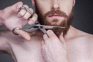 Cutting his perfect beard. Close-up of handsome young shirtless man cutting his beard with scissors while standing against grey background photo