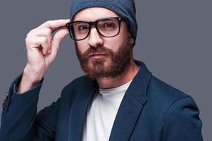 In my own style. Handsome young bearded man in smart casual wear adjusting his eyeglasses and looking at camera while standing against grey background photo