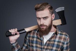 Confident lumberjack. Confident young bearded man carrying a big axe on shoulder and looking at camera while standing against grey background photo